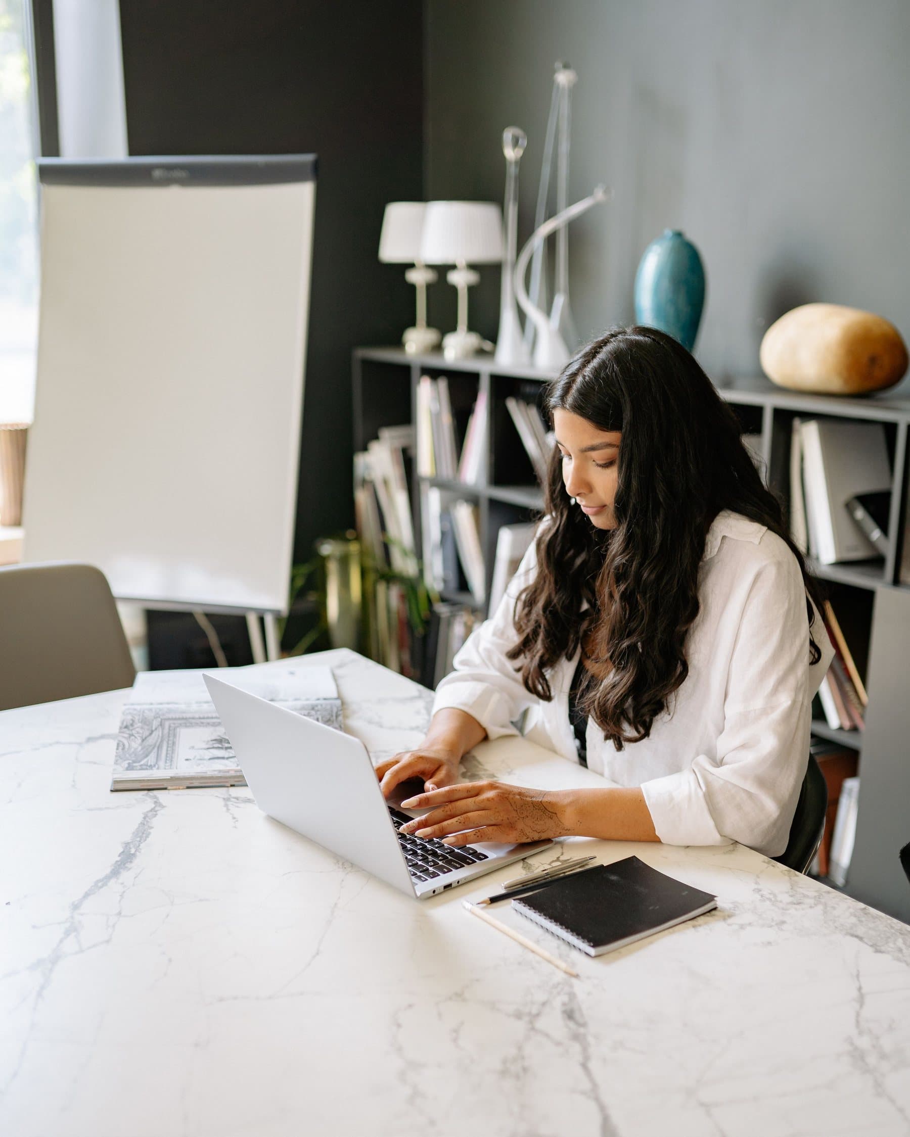 A woman works on her laptop at a marble table beside lamps and bookshelves