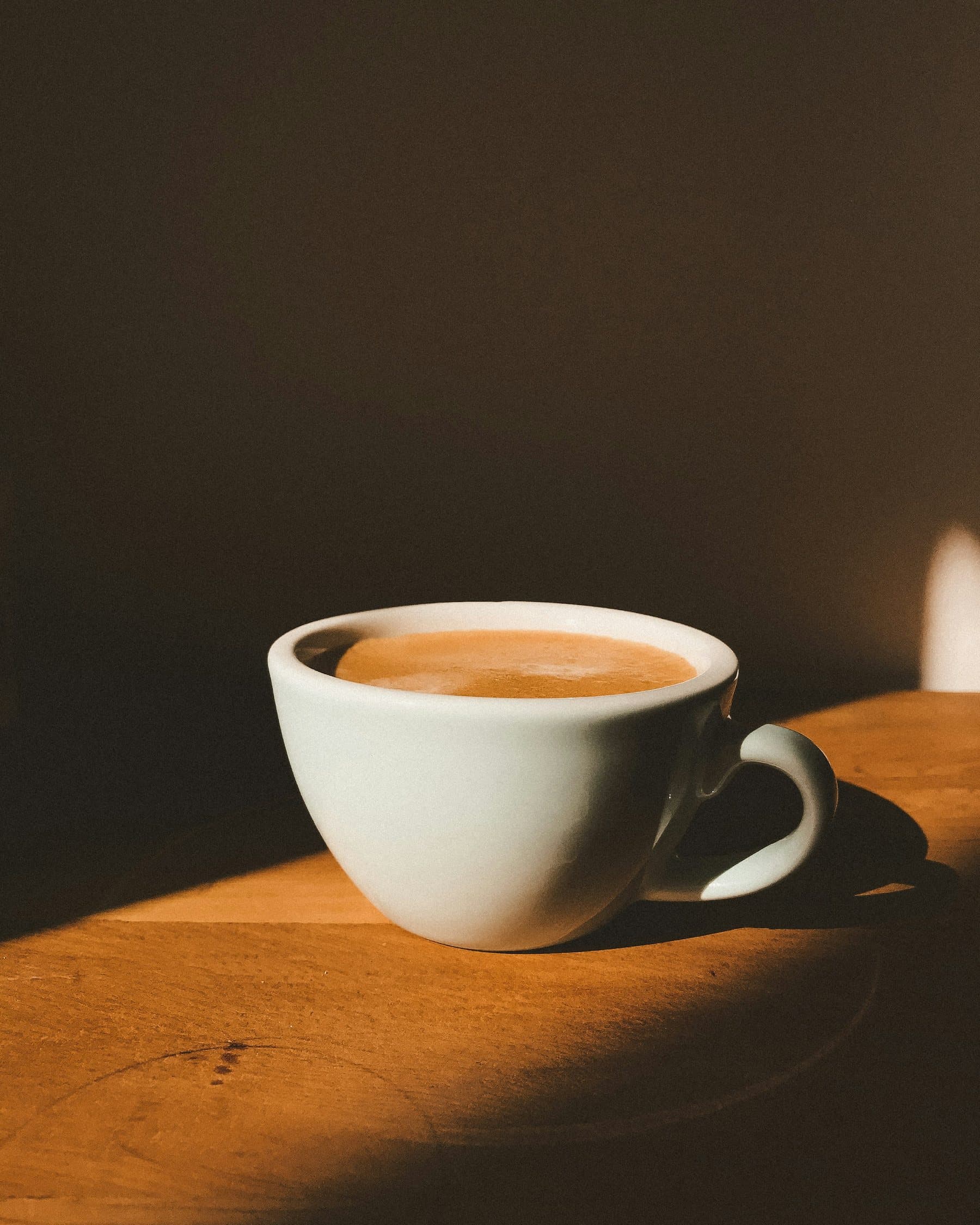 White cup of coffee on a wooden table catching a band of golden sunlight