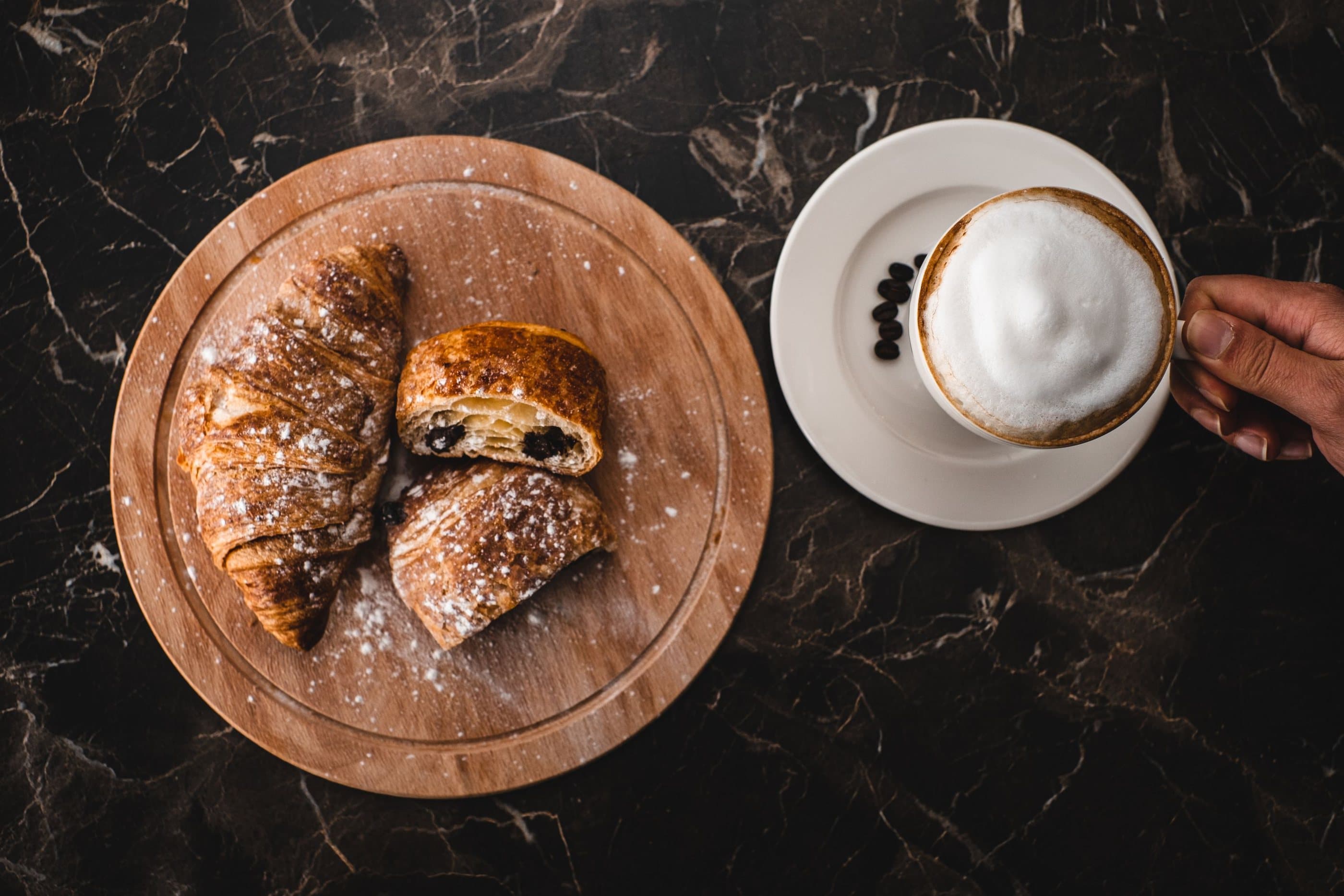 Croissants on a wooden board beside a cappuccino on a dark marble table, seen from above