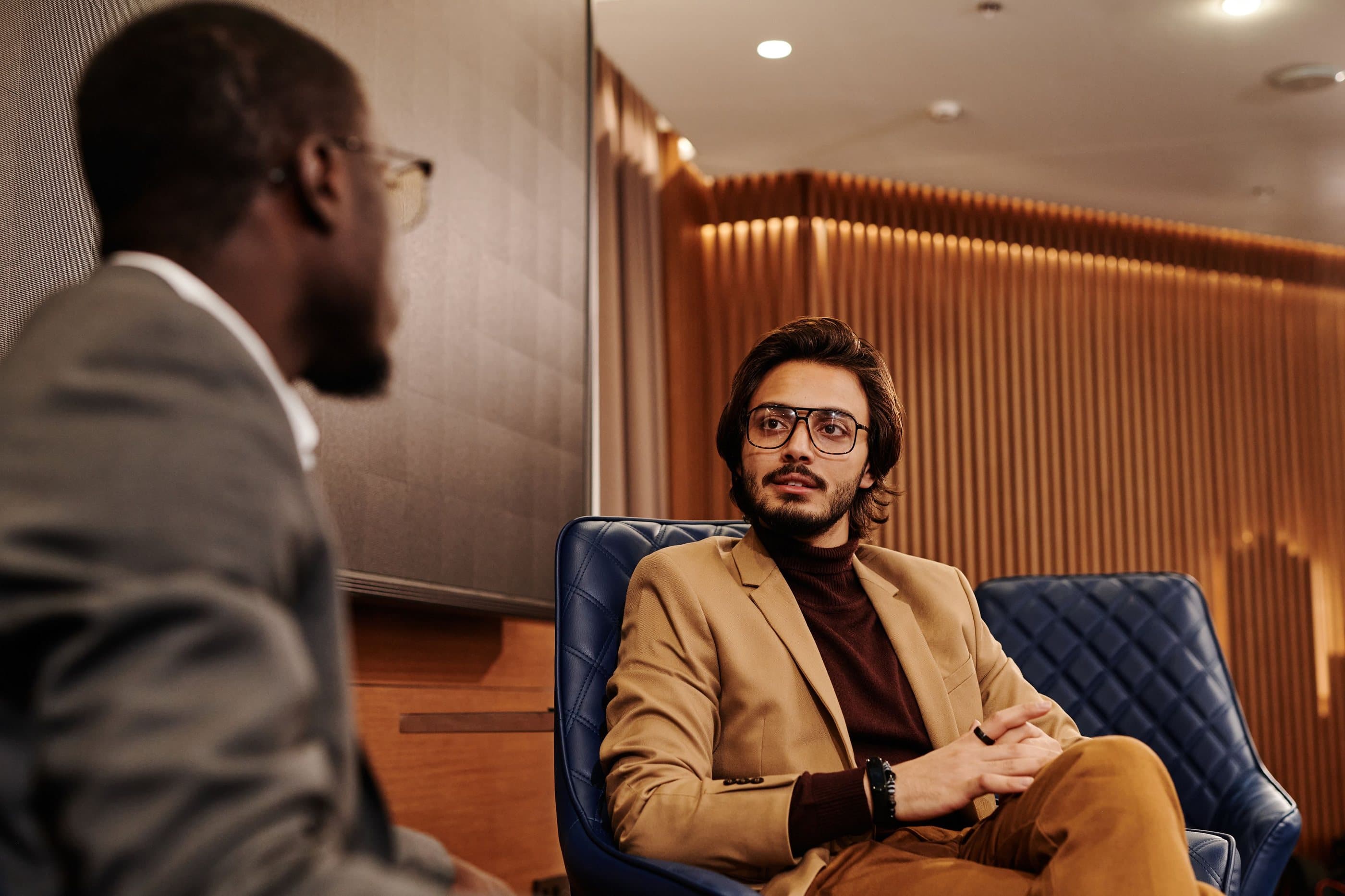 A professional in a camel blazer listens in a quilted leather armchair during a private conversation in a wood-slatted lounge