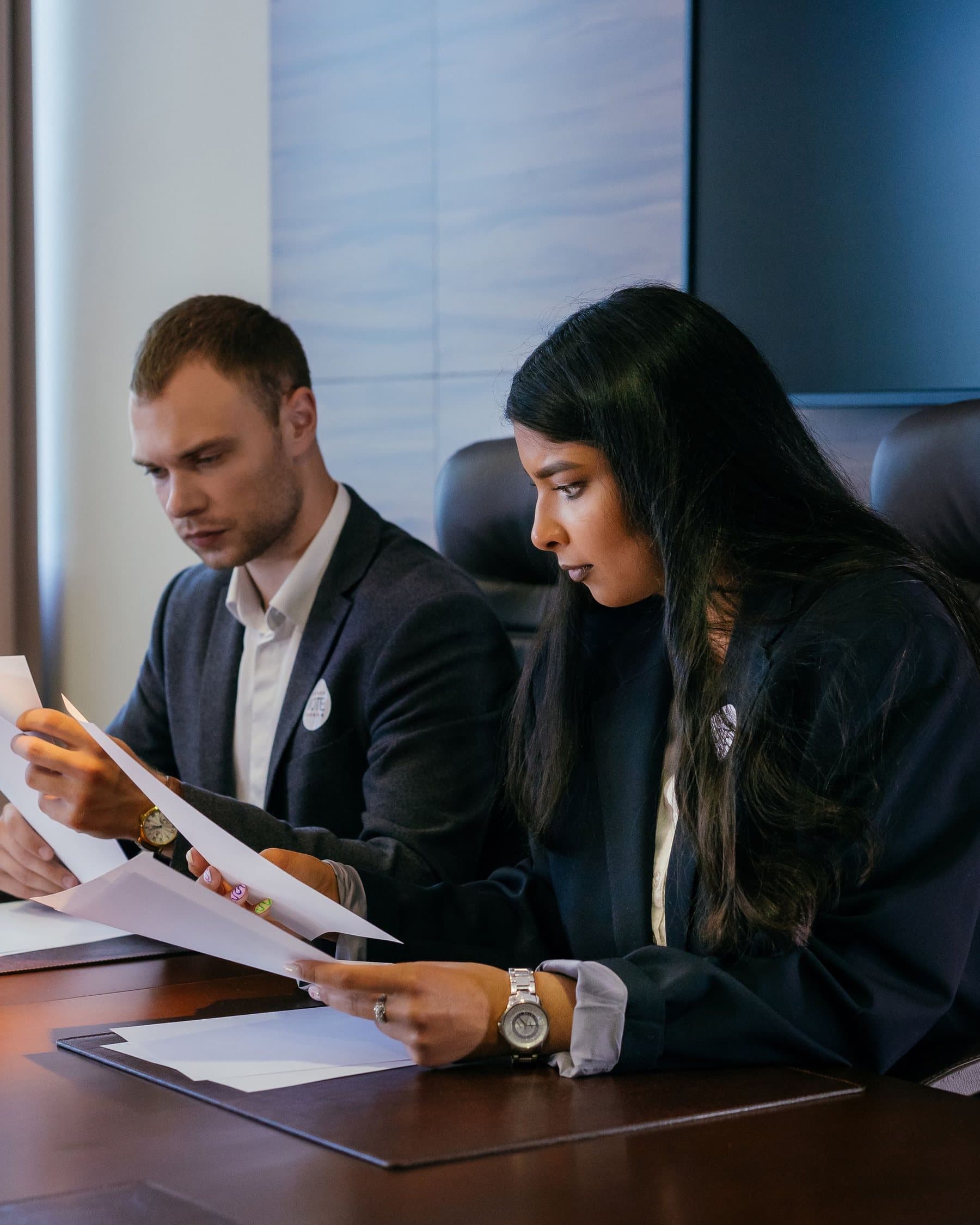 Two colleagues in dark suits reviewing printed documents together at a polished boardroom table