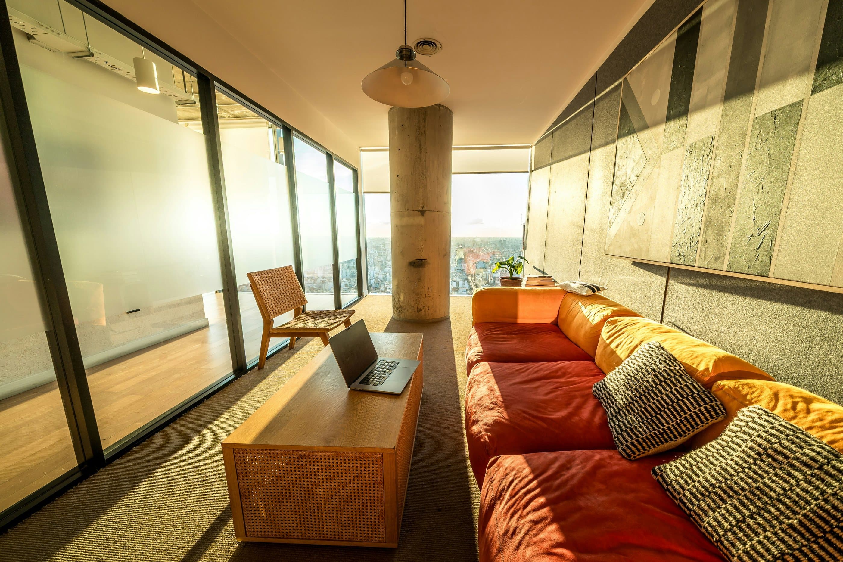 Sunlit lounge corridor with a long terracotta sofa, woven cushions and a laptop resting on a low wooden bench beside floor-to-ceiling glass