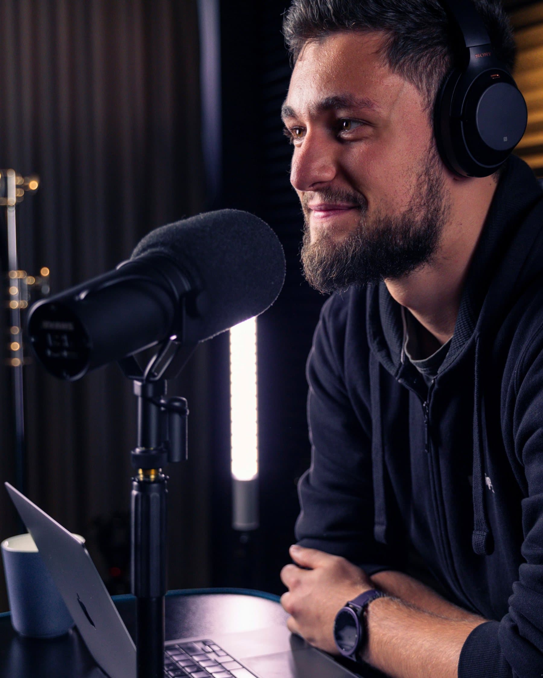 A host in headphones smiles into a broadcast microphone inside a warm, acoustically treated recording booth