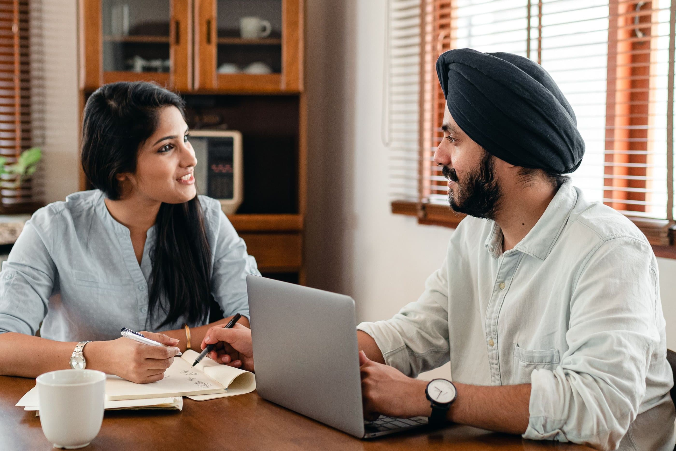 A woman and a Sikh man talk over a laptop and notebook at a warm wooden table