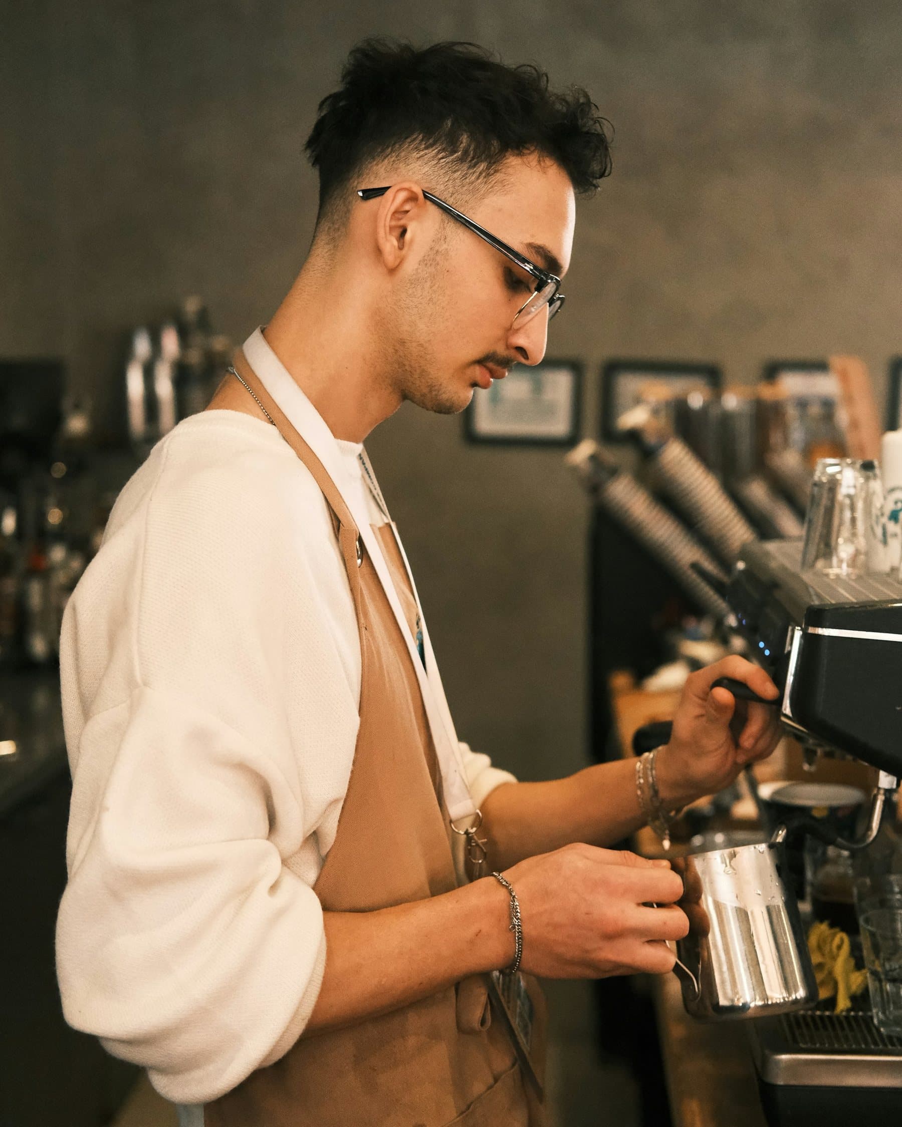 Barista in a tan apron steaming milk at an espresso machine