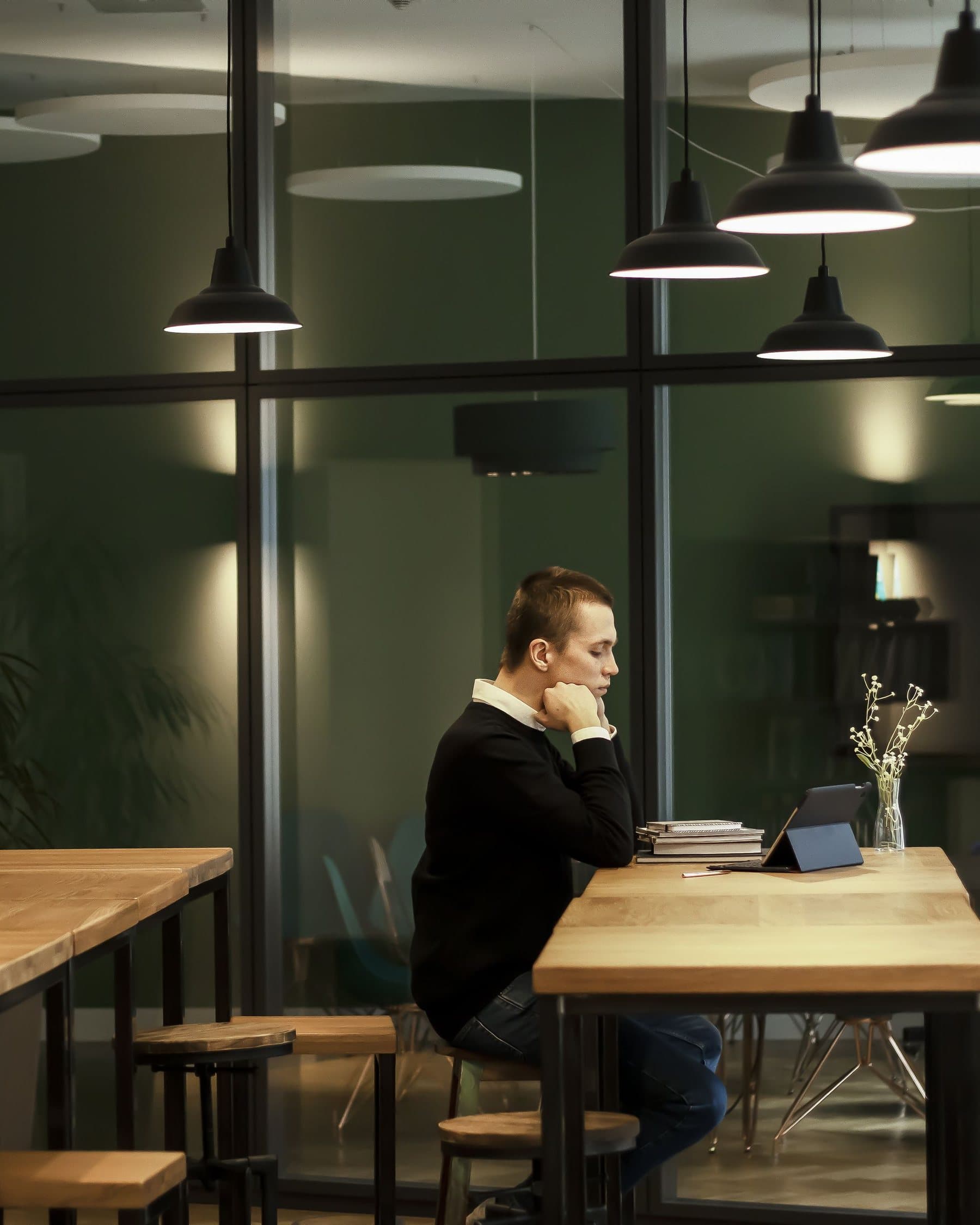 A professional working quietly at a long oak table under low pendant lights, with a tablet, books and a small vase of flowers