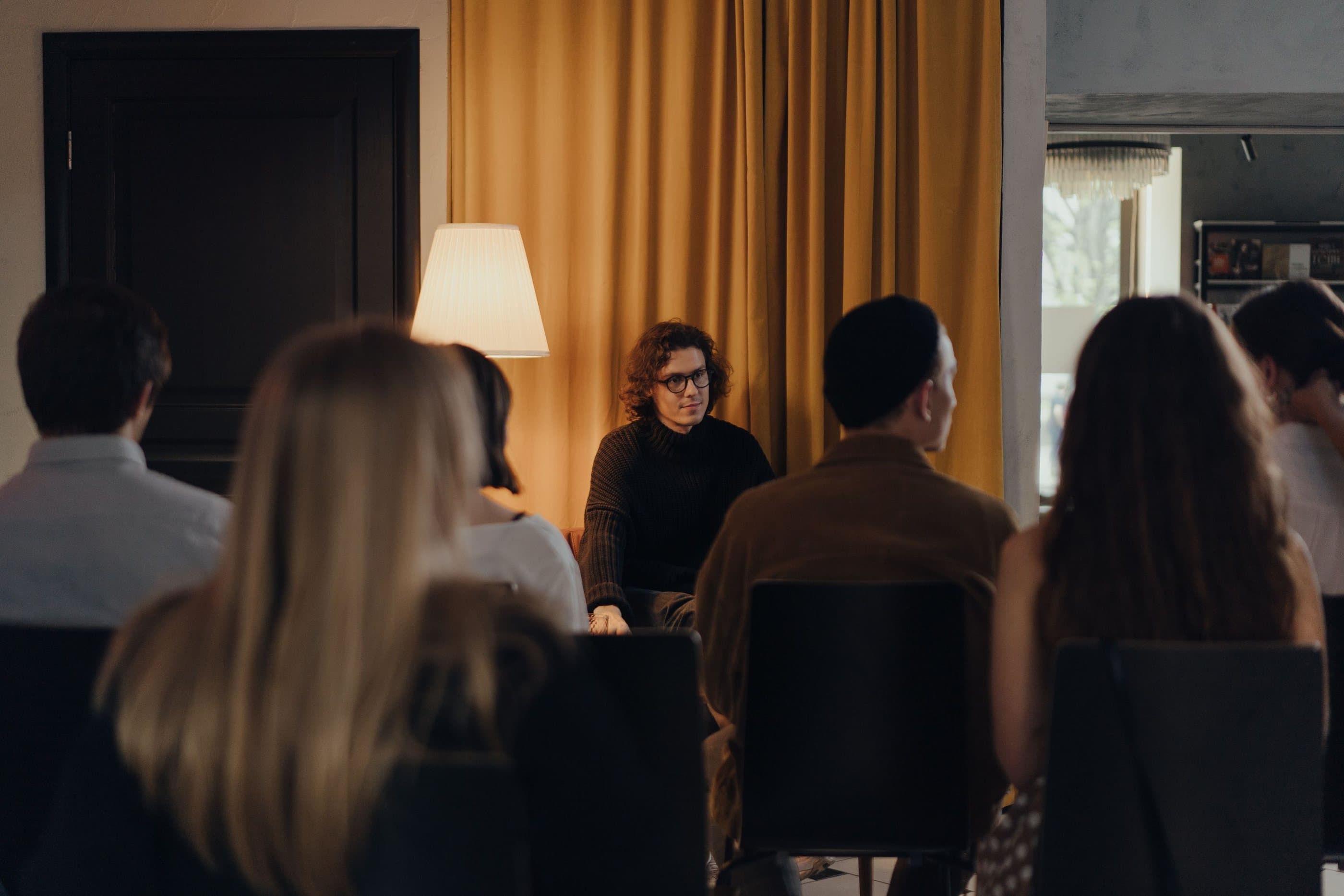 A small audience listens to a speaker in a warm room with amber curtains and a floor lamp